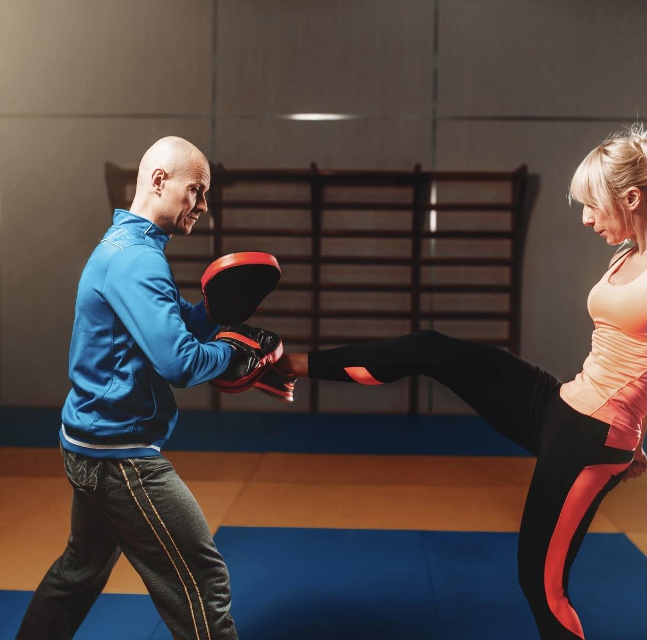 Trainer hält Pratzen, während eine Sportlerin einen Kick ausführt. Beide befinden sich in einem Trainingsraum.