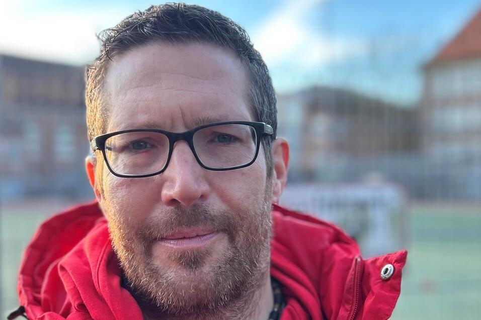 Mann mit Brille in roter Jacke und Sweatshirt, steht im Freien vor einem Sportfeld.