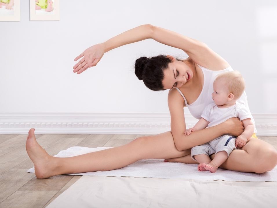 Mutter sitzt auf dem Boden und streckt sich, während sie ihr Baby auf dem Schoß hält. Helle, freundliche Raumgestaltung.