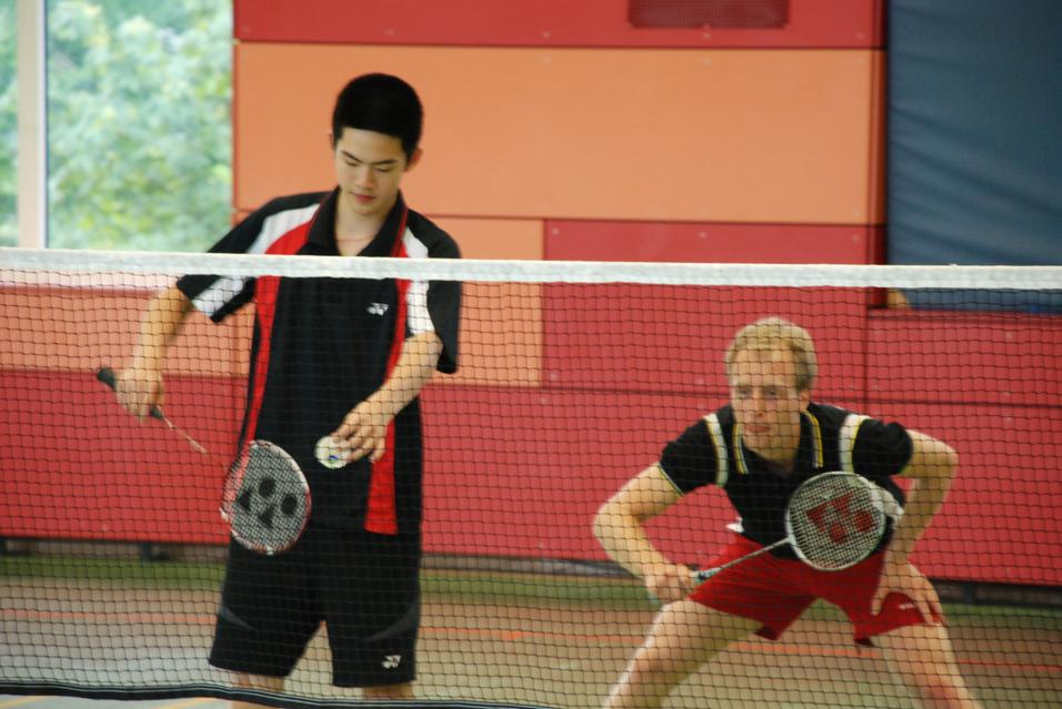 Zwei Badmintonspieler in einer Halle stehen sich am Netz gegenüber, bereit zum Spiel mit Schlägern in der Hand.