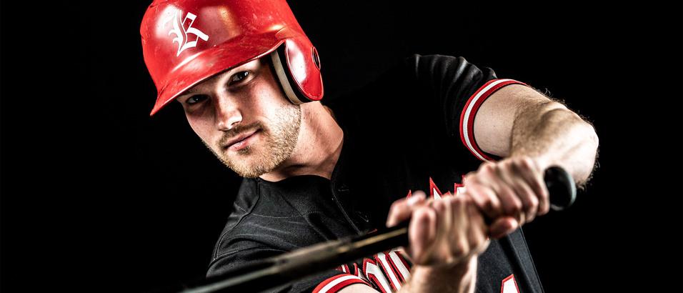Ein Spieler in rotem Helm und schwarzem Trikot hält einen Baseballschläger und blickt konzentriert zur Kamera.