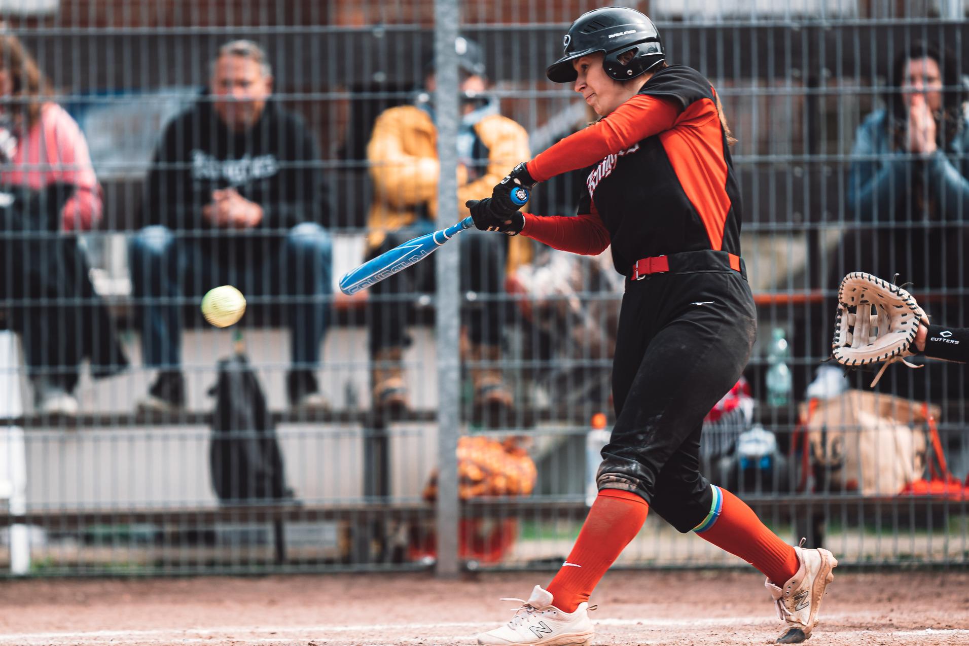 Softballspielerin schlägt einen Ball mit ihrem Schläger, während Zuschauer im Hintergrund auf einer Tribüne sitzen.