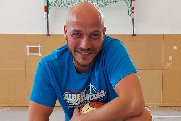 Mann in blauer Sportkleidung kniet umgeben von Basketballen in einer Sporthalle.