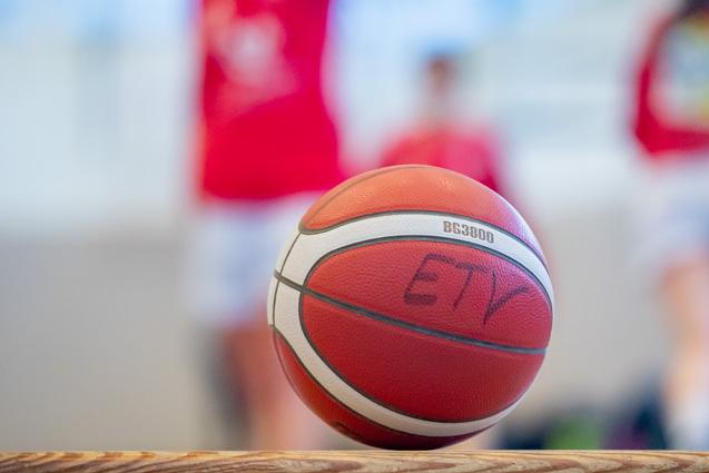 Nahaufnahme eines Basketballs mit einer Aufschrift, unscharfer Hintergrund von Spielern in roten Trikots.