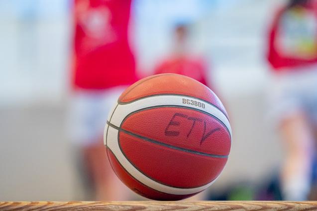 Nahaufnahme eines Basketballs mit einer Aufschrift, unscharfer Hintergrund von Spielern in roten Trikots.