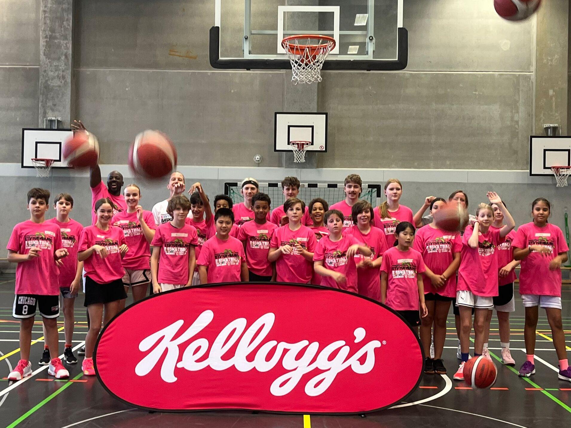 Gruppe von Kindern in pinken T-Shirts steht vor einem Basketballkorb, während sie Bälle in die Luft werfen.