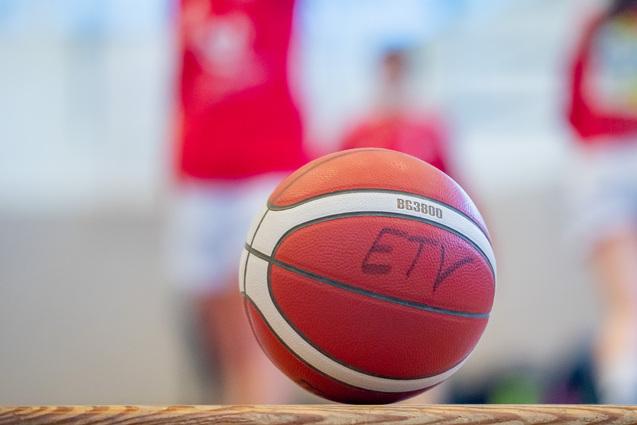 Nahaufnahme eines Basketballs mit einer Aufschrift, unscharfer Hintergrund von Spielern in roten Trikots.