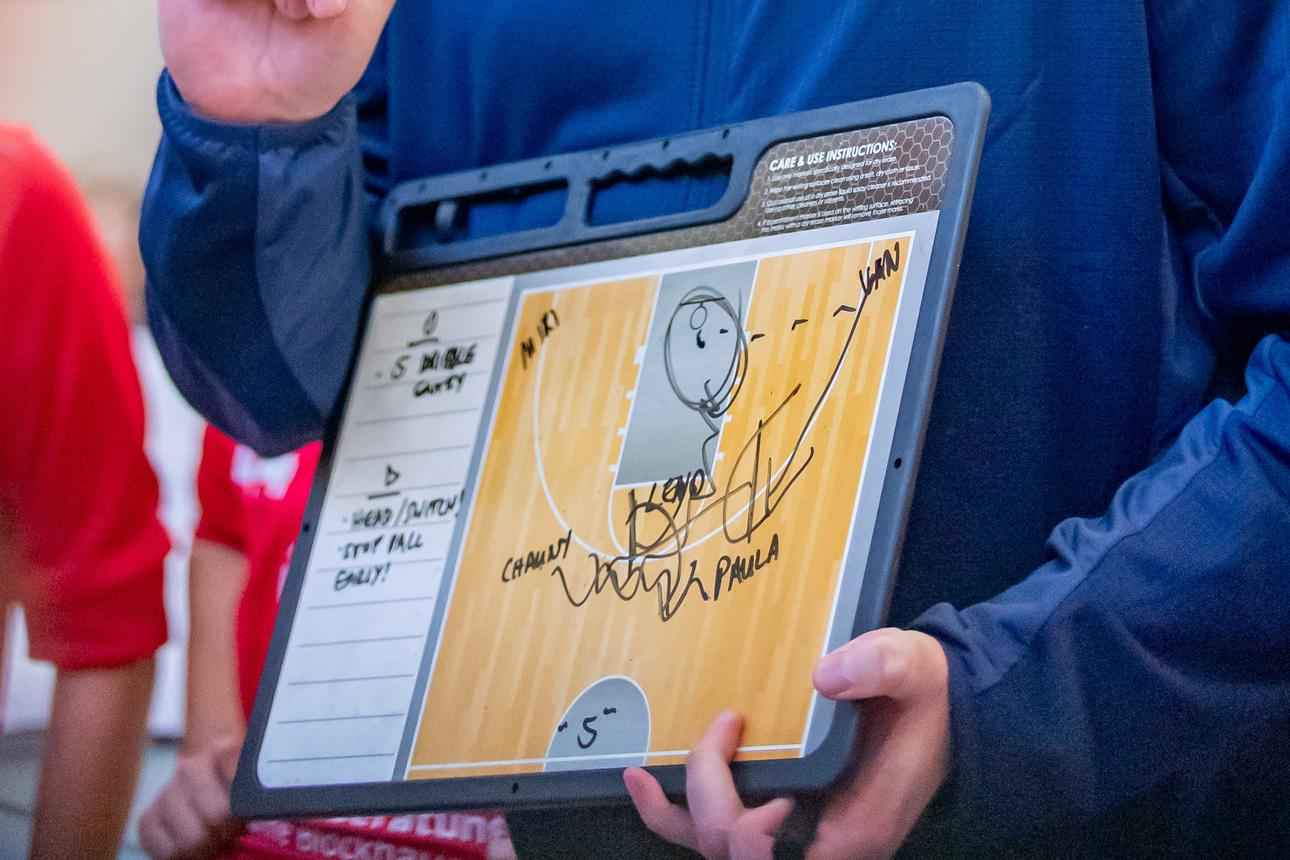 Trainer hält ein Taktikboard mit Zeichnungen und Notizen, während er mit einer Gruppe von Spielern spricht.