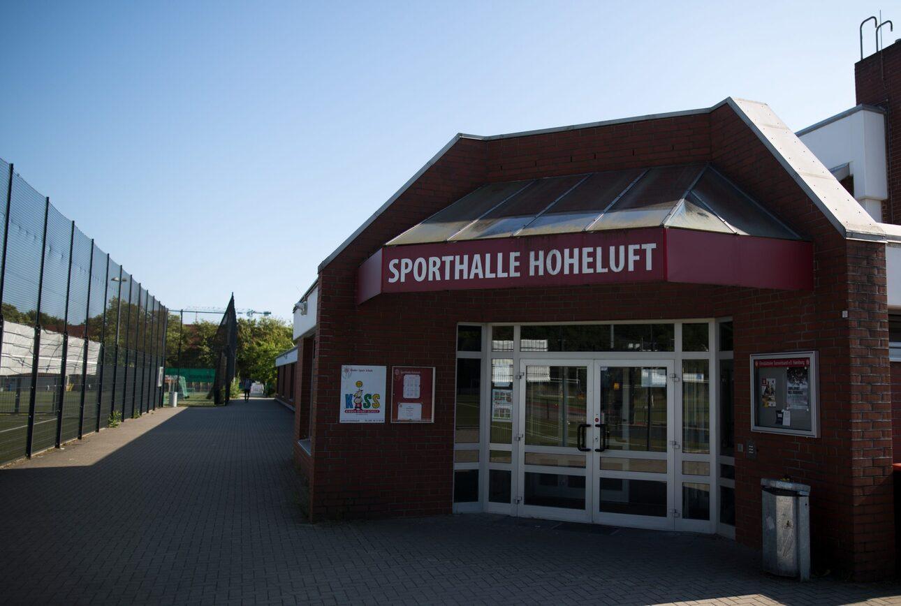 Eingang der Sporthalle Hoheluft mit angrenzendem Außenbereich und Tennisplätzen im Hintergrund an einem sonnigen Tag.
