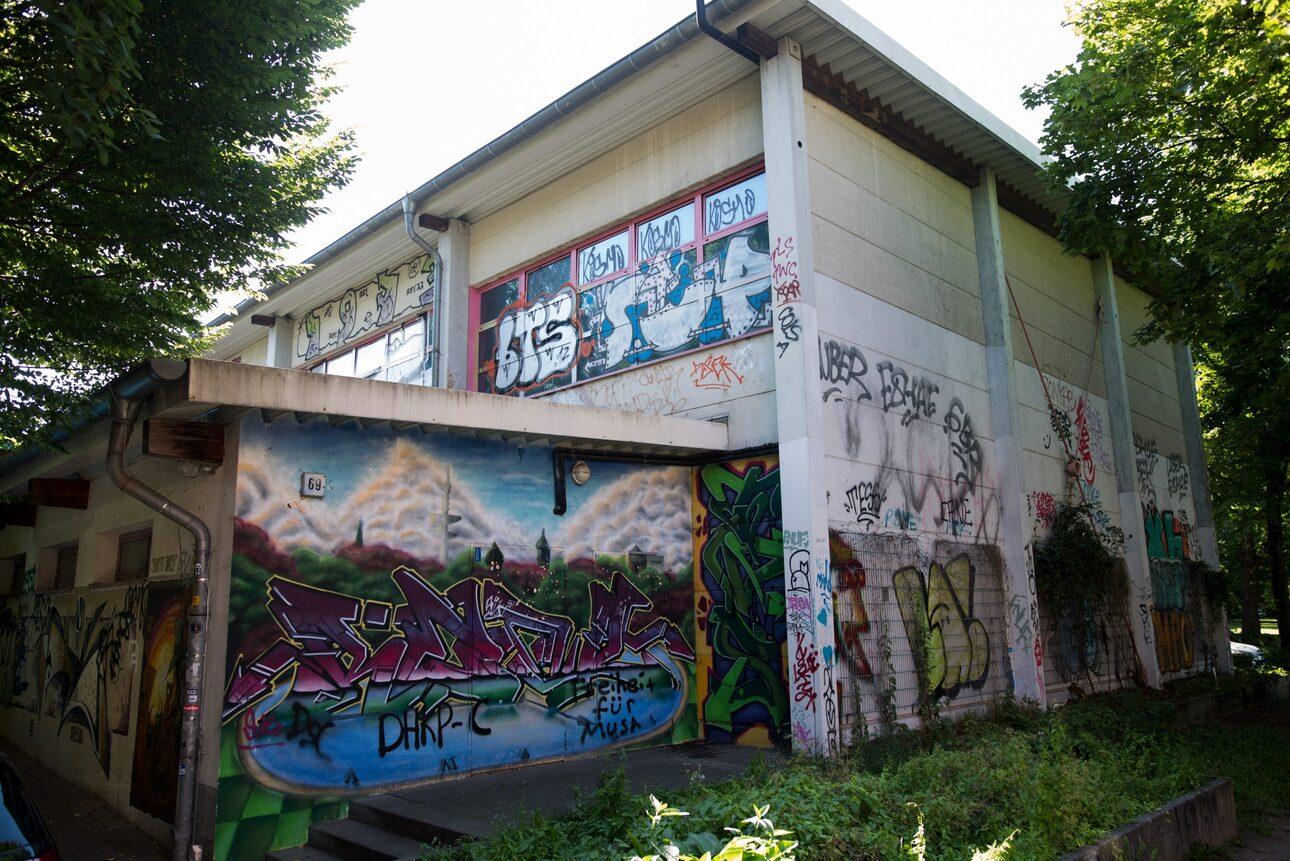 Graffiti-bedeckte Außenwand eines Gebäudes umgeben von Bäumen und grünem Gras im Tageslicht.