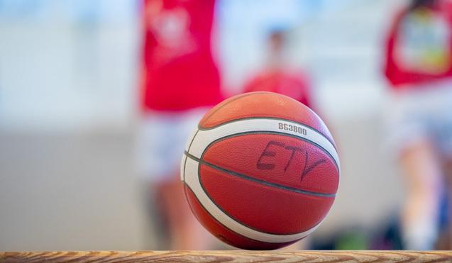 Nahaufnahme eines Basketballs mit einer Aufschrift, unscharfer Hintergrund von Spielern in roten Trikots.