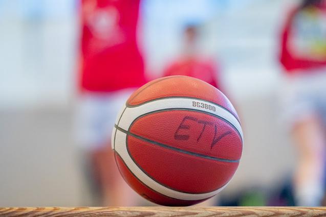 Nahaufnahme eines Basketballs mit einer Aufschrift, unscharfer Hintergrund von Spielern in roten Trikots.