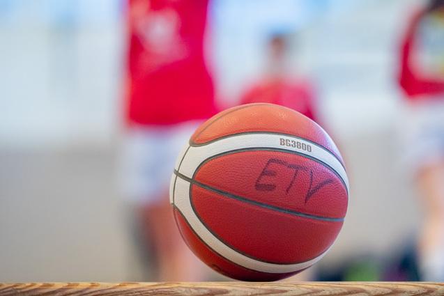 Nahaufnahme eines Basketballs mit einer Aufschrift, unscharfer Hintergrund von Spielern in roten Trikots.