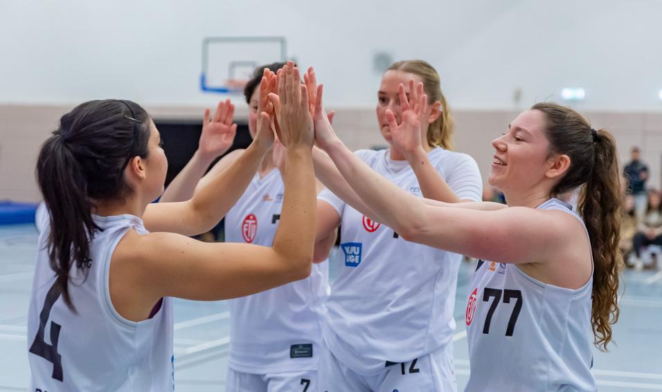 Vierte Frauen im weißen Basketball-Trikot feiern mit einem High-Five in einer Sporthalle.