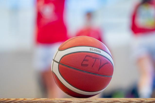 Nahaufnahme eines Basketballs mit einer Aufschrift, unscharfer Hintergrund von Spielern in roten Trikots.