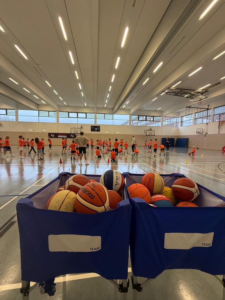 Zwei Basketballkörbe mit Bällen im Vordergrund, dahinter Kinder in orangefarbenen Shirts beim Training in einer Halle.