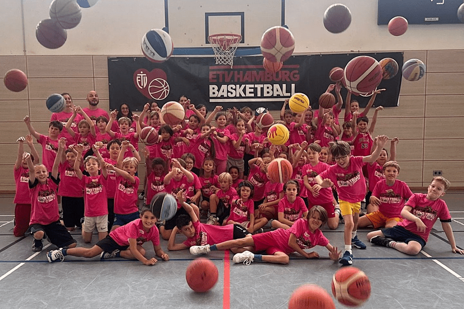Gruppe von Jungen und Mädchen in pinken T-Shirts posiert auf einem Basketballplatz, umringt von schwebenden Bällen.