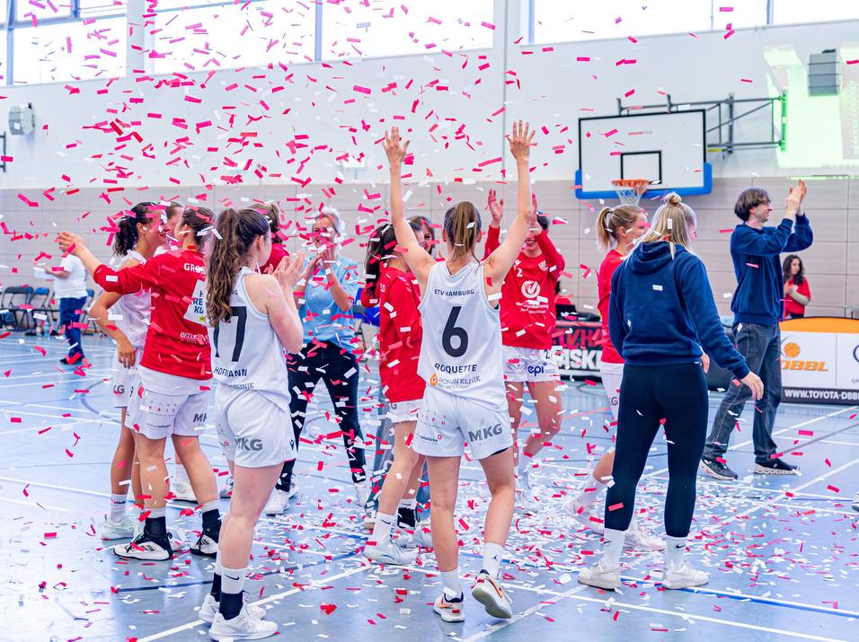 Basketballmannschaft feiert mit Konfetti auf dem Spielfeld nach einem gewonnenen Spiel. Spielerinnen jubeln und applaudieren.