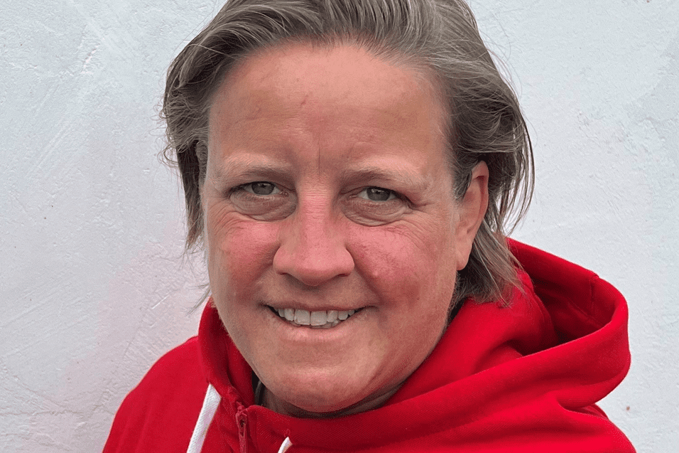 Person mit kurzen Haaren in rotem Hoodie mit Basketball-Logo vor weißer Wand. Lächelt freundlich in die Kamera.