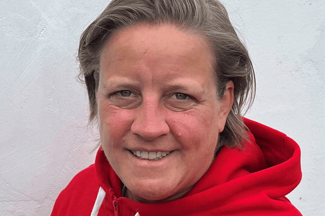 Person mit kurzen Haaren in rotem Hoodie mit Basketball-Logo vor weißer Wand. Lächelt freundlich in die Kamera.