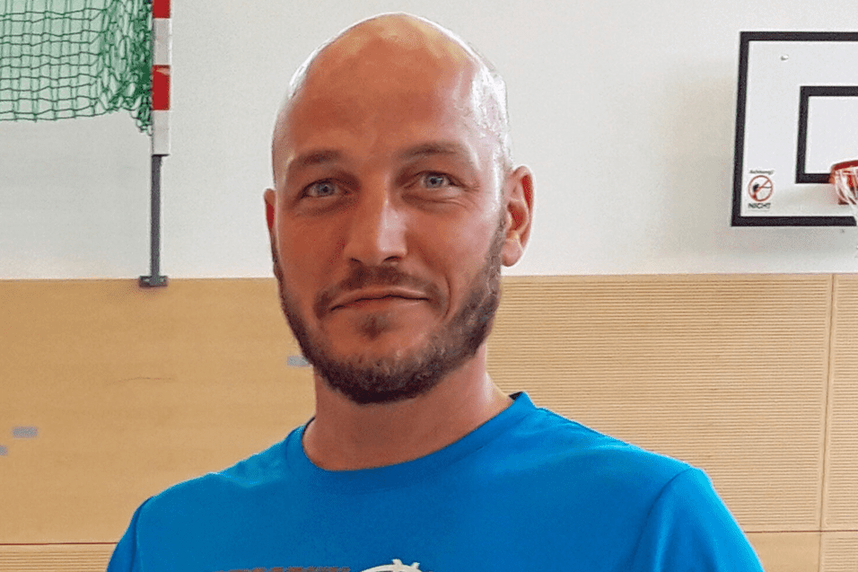 Mann in blauer Sportkleidung hält einen Basketball, steht in einer Sporthalle mit Basketballkorb im Hintergrund.