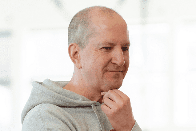 Nachdenklicher Mann in grauem Hoodie, der mit Hand an Kinn in eine Richtung blickt, vor heller Hintergrundumgebung.