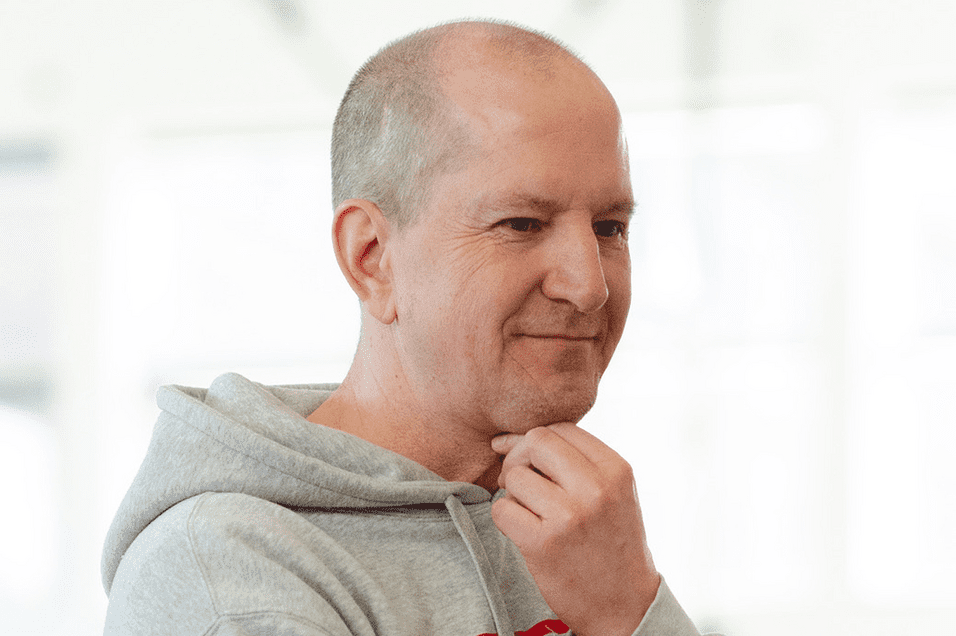 Nachdenklicher Mann in grauem Hoodie, der mit Hand an Kinn in eine Richtung blickt, vor heller Hintergrundumgebung.