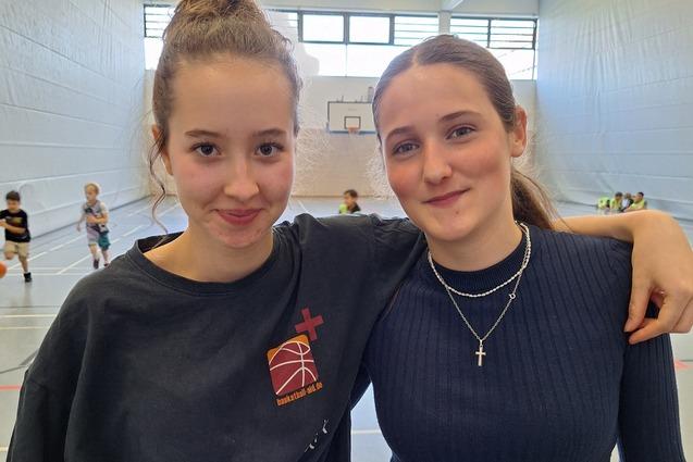 Zwei Mädchen posieren lächelnd nebeneinander in einer Sporthalle mit Basketballkorb im Hintergrund.