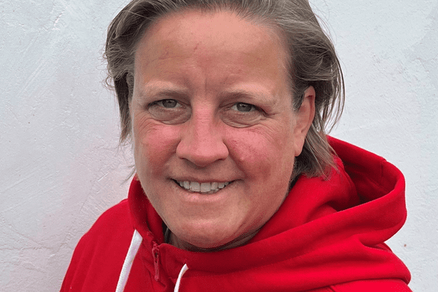 Person mit kurzen Haaren in rotem Hoodie mit Basketball-Logo vor weißer Wand. Lächelt freundlich in die Kamera.