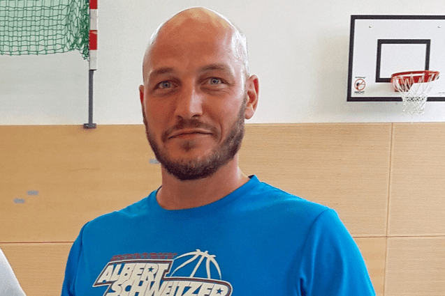 Mann in blauer Sportkleidung hält einen Basketball, steht in einer Sporthalle mit Basketballkorb im Hintergrund.