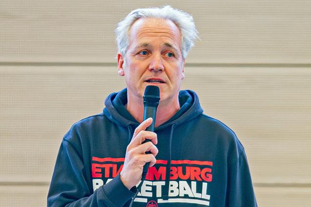 Mann mit Mikrofon hält eine Rede, trägt einen Hoodie mit dem Aufdruck „ETTILM BURG BASKETBALL“.