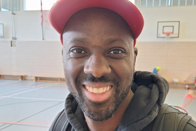 Lächelnder Mann mit schwarzem Hoodie und roter Cap steht in einer Sporthalle mit Spielern im Hintergrund.