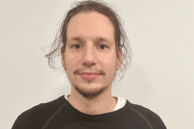 Portrait eines Mannes mit mittellangen Haaren, der ein schwarzes Basketball-Shirt mit Aufschrift trägt. Hintergrund ist weiß.