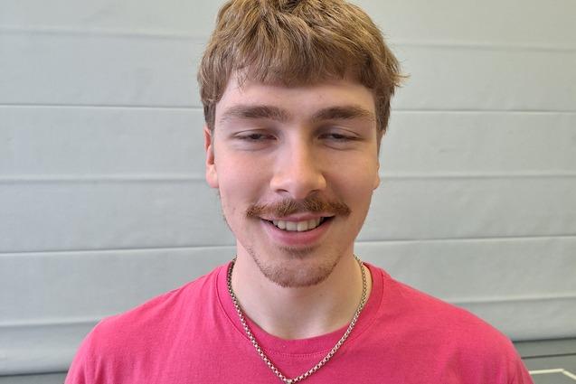 Junger Mann mit kurzem, lockigem Haar, lächelt in einem rosa T-Shirt mit Basketballmotiv, vor einer grauen Wand.