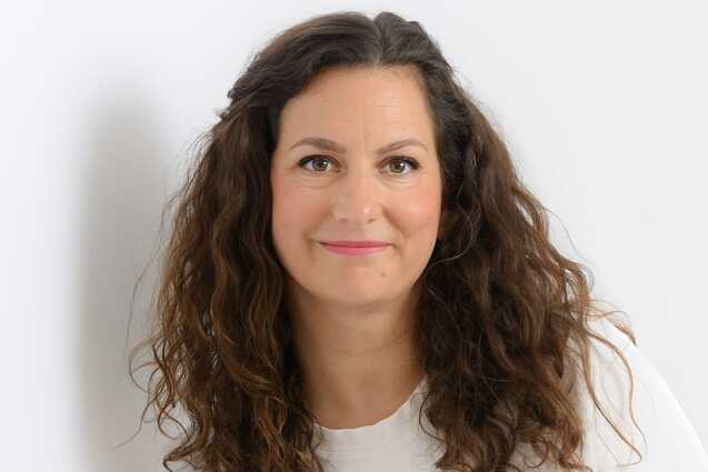 Porträt einer lächelnden Frau mit lockigem Haar, die in einem weißen Oberteil vor einem hellen Hintergrund steht.