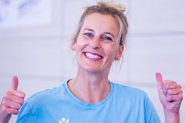 Lächelnde Frau in blauer T-Shirt, zeigt Daumen hoch vor neutralem Hintergrund.