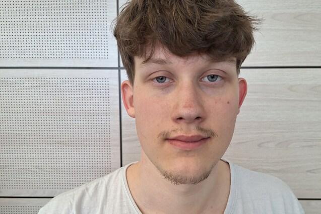 Porträt eines jungen Mannes mit kurzem, lockigem Haar, der in einem hellen T-Shirt vor einer Holzpaneelwand steht.