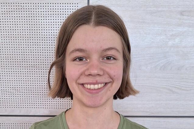 Lächelnde Person mit kurzem Haar in einem grünen Nike-Shirt, steht vor einer Holzpaneel-Wand.