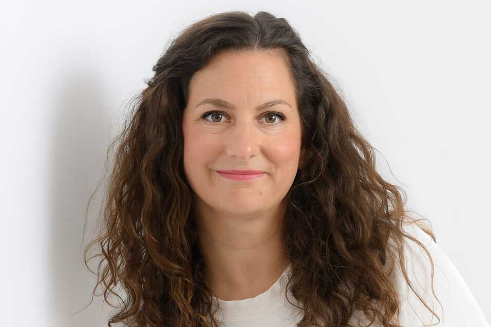 Porträt einer lächelnden Frau mit lockigem Haar, die in einem weißen Oberteil vor einem hellen Hintergrund steht.