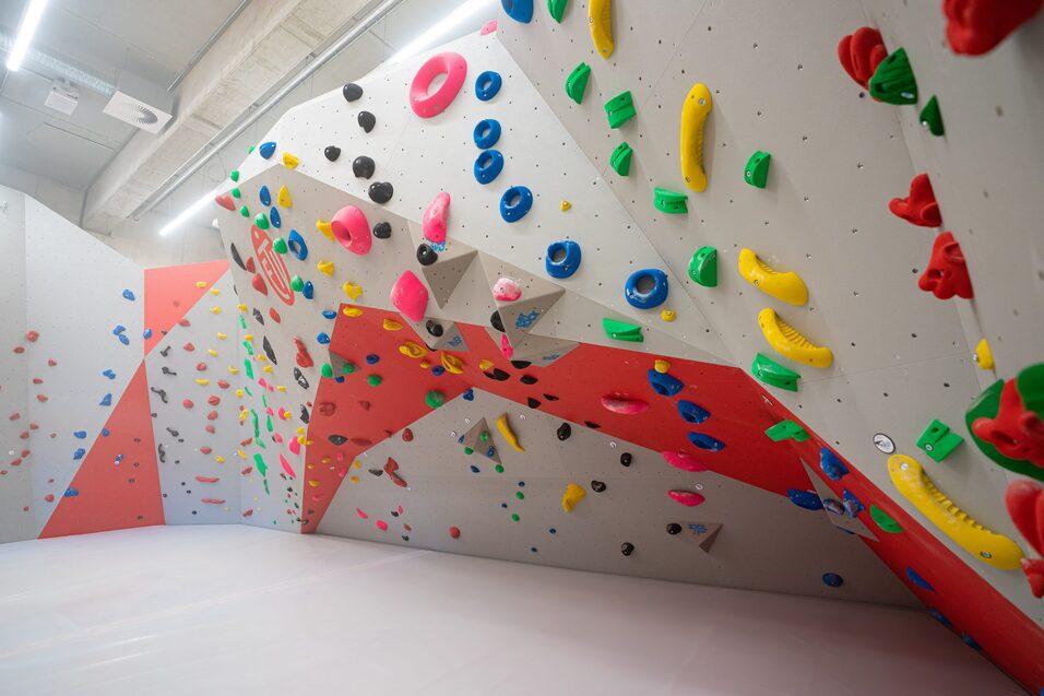 Boulderwand mit bunten Griffen in einem Kletterraum, im Hintergrund eine großzügige Fläche zum Klettern.