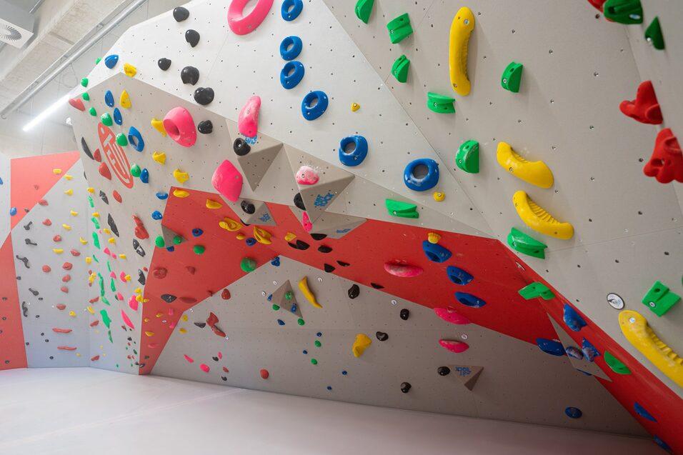Boulderwand mit bunten Griffen in einem Kletterraum, im Hintergrund eine großzügige Fläche zum Klettern.