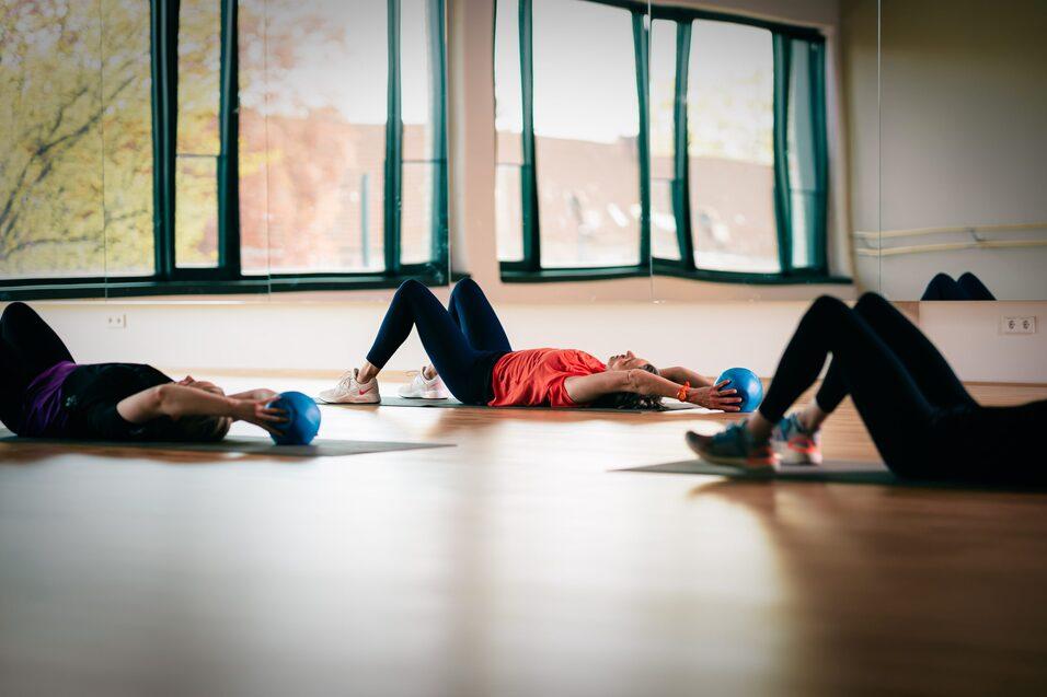 Drei Personen liegen auf Matten in einem Fitnessraum und halten jeweils einen kleinen Ball in der Hand.