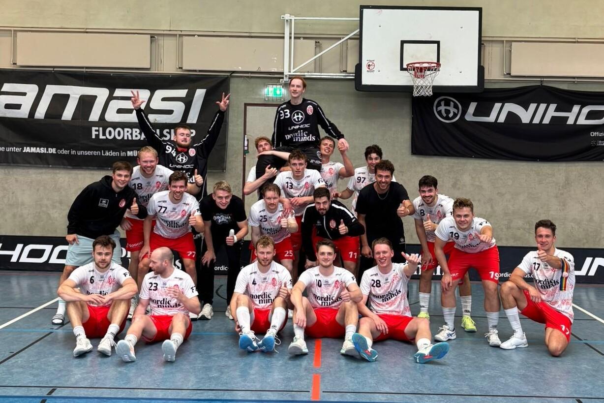Gruppenfoto von 17 Sportlern in roten und weißen Trikots auf einem Sportboden, umgeben von Werbebannern und einer Anzeigetafel.