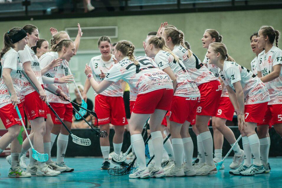 Feiernde Mannschaft von Spielerinnen in roten Shorts und weißen Trikots versammelt auf dem Sportboden, bereit fürs Spiel.
