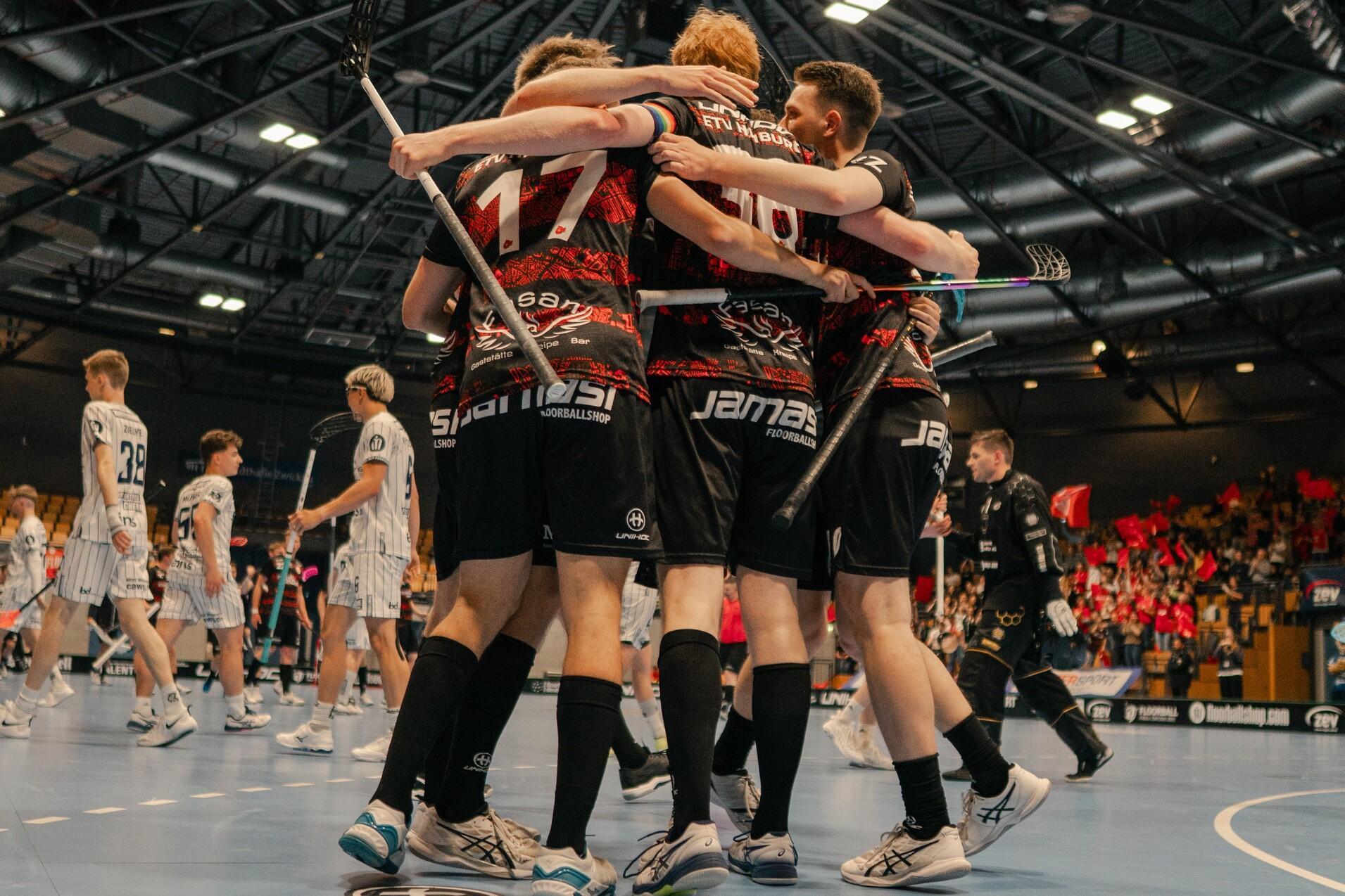 Gruppenumarmung von Spielern in schwarz-roten Trikots auf einem Hallenboden während eines Unihockeyspiels.