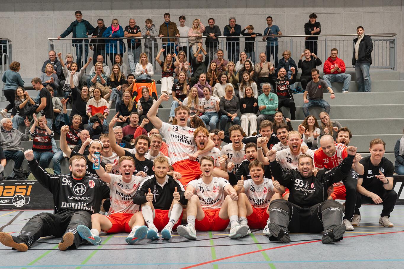 Gruppenselfie einer Handballmannschaft mit jubelnden Spielern und Zuschauern in einer Sporthalle.