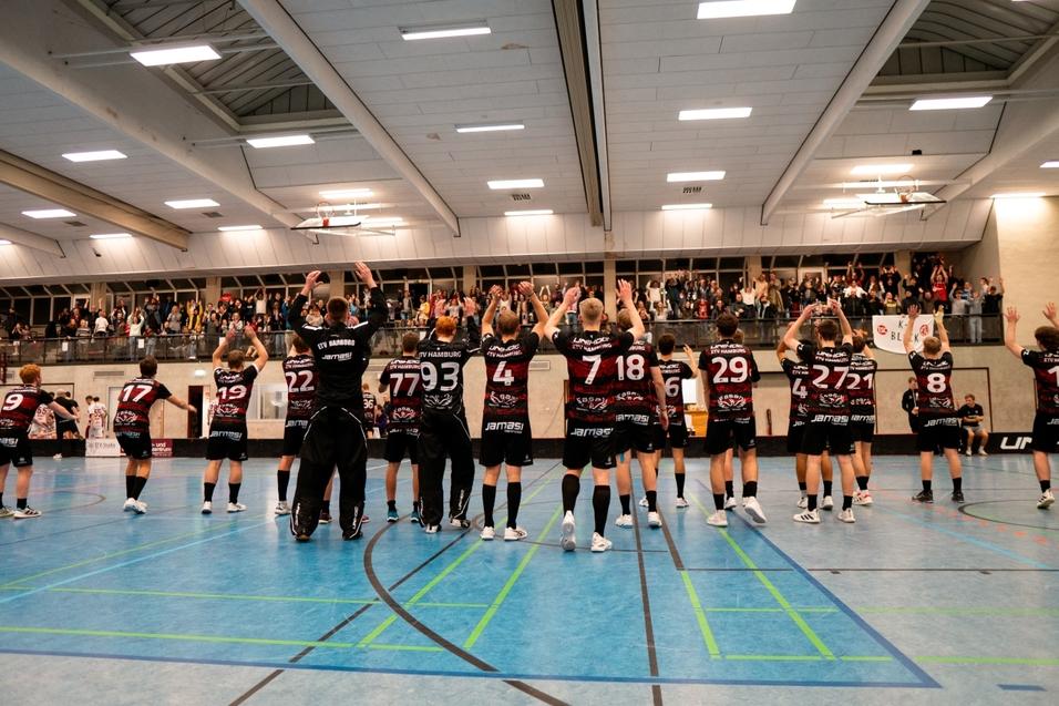 Mannschaft in schwarzen Trikots feiert im Sporthall mit erhobenen Händen vor jubelndem Publikum.