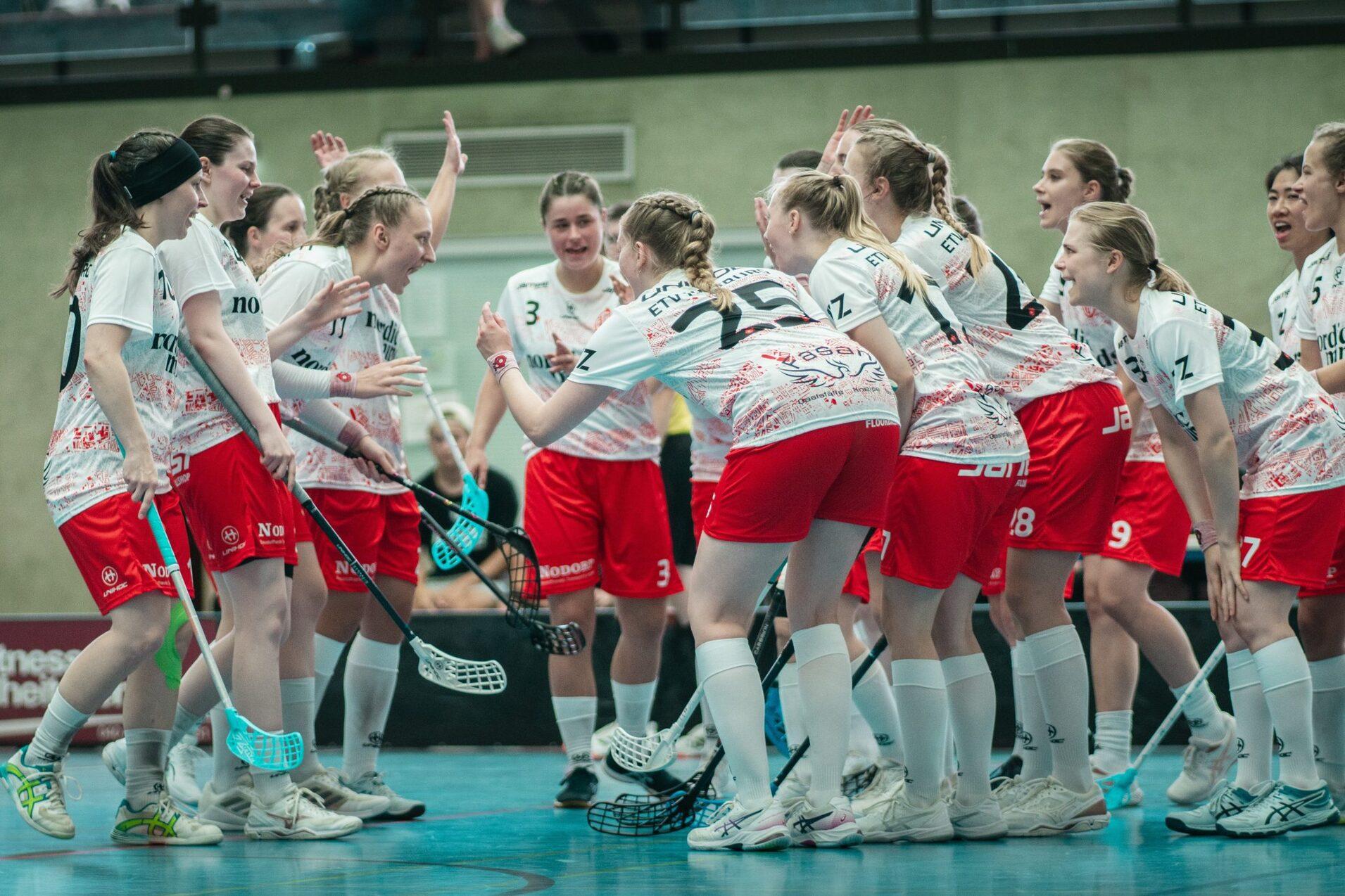 Feiernde Mannschaft von Spielerinnen in roten Shorts und weißen Trikots versammelt auf dem Sportboden, bereit fürs Spiel.