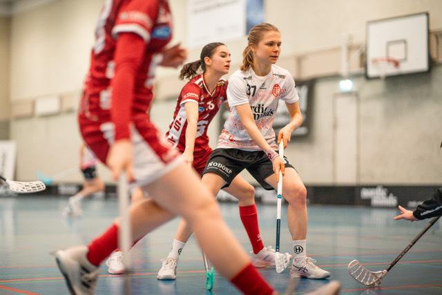 Aktion auf dem Feld mit zwei Spielerinnen in roten und weißen Trikots beim Unihockeyspiel in einer Sporthalle.