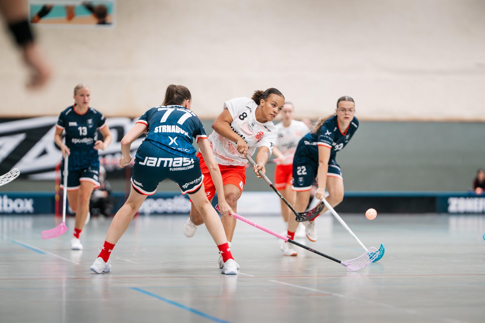 Zwei Gruppen von Spielerinnen im Floorball kämpfen um den Ball in einer Sporthalle, eine Spielerin hat den Schläger erhoben.